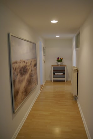 Een hal met een houten vloer, een groot schilderij aan de linkerkant, een kleine tafel met een bloevas en een radiator aan de rechterkant.
