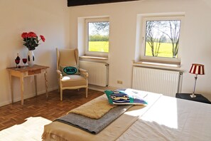 Een slaapkamer met een bed, een stoel, een tafel met een bloevas en twee ramen met uitzicht op een veld.