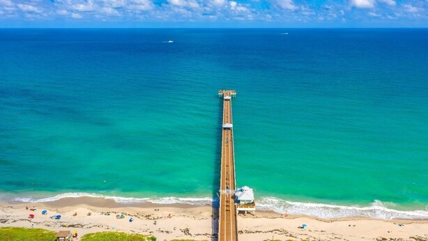 Eine Steg, der sich in türkisfarbenes Wasser erstreckt, mit einem Strand und Sonnenschirmen am Ende.