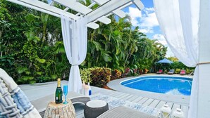 Ein Poolbereich mit Holzterrasse, Liegestühlen, einem kleinen Tisch mit Getränken und einer weißen Markise.