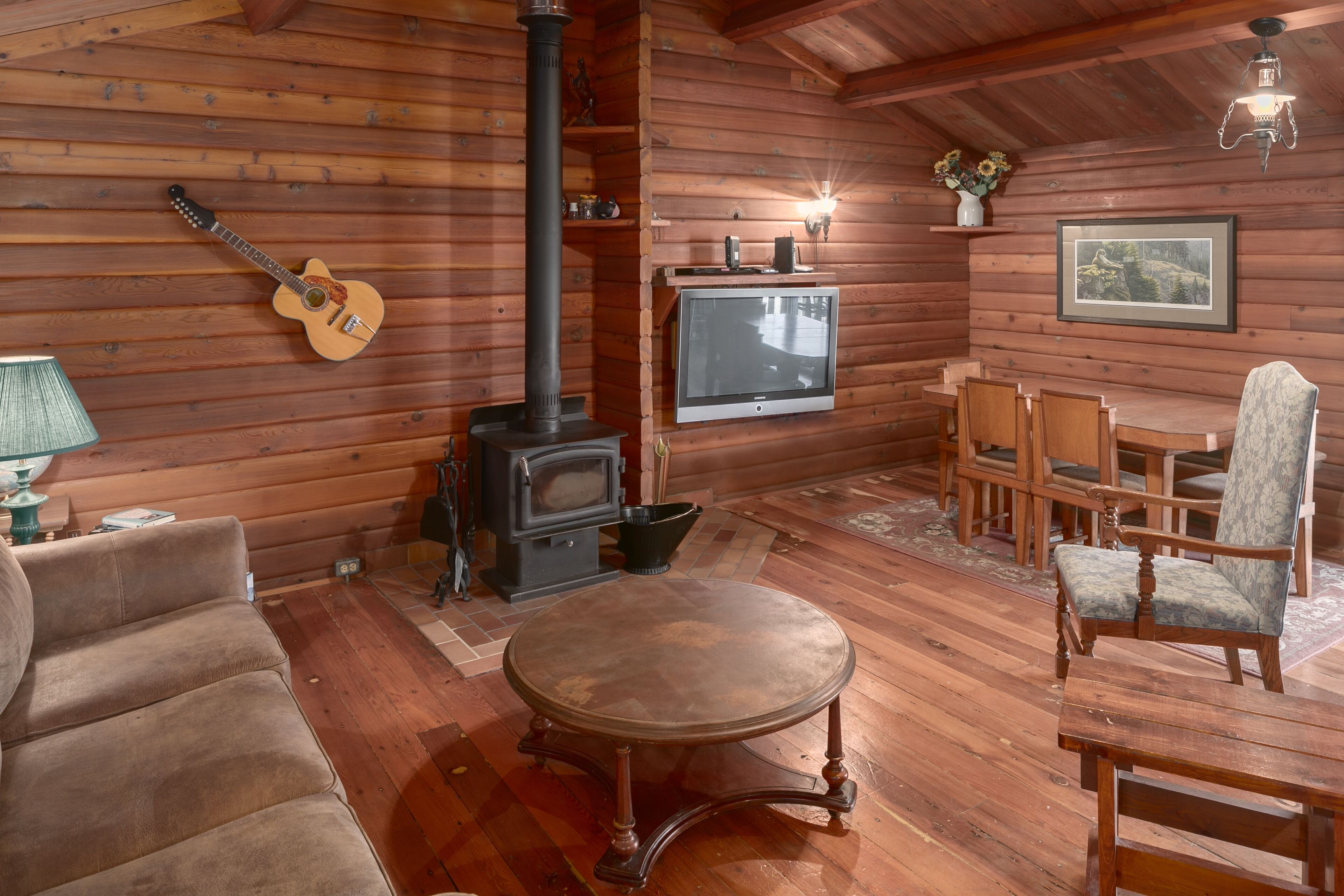 A cozy cabin interior with a wooden floor, a fireplace, a television, and a dining area with chairs and a table.