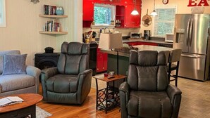 Una sala moderna con sillones de cuero, un sofá y una mesa de centro. Se ve una cocina al fondo con armarios rojos y electrodomésticos de acero inoxidable.