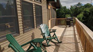 A wooden deck with Adirondack chairs, a barbecue grill, and a view of a forested area.