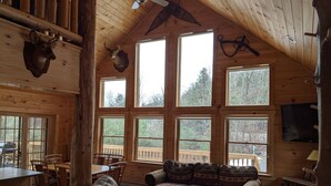 A cozy cabin interior with a wooden ceiling, large windows, a dining area, and a sofa.