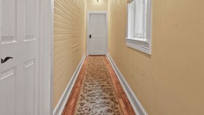 A hallway with wooden flooring, a patterned rug, a white door, and a window with a blind.