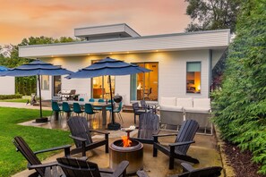 Ein modernes Haus mit einer Terrasse, die Gartenmöbel, eine Feuerstelle und Sonnenschirme bietet.