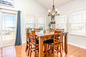 A dining room with a wooden table and chairs, large windows with blinds, a chandelier, and a view of the ocean. - Beaches B Tripp'n - Awesome 4 BR Home - Ocean View and Elevator! (North Topsail Beach)