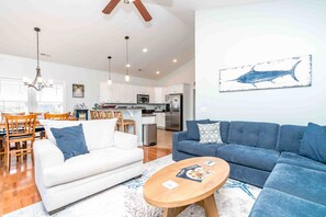 A modern living room with a white sofa, a blue sofa, and a wooden coffee table. There is a kitchen in the background with white cabinets and a refrigerator.