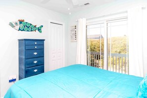 A bedroom with a blue bedspread, a blue dresser with drawers, a white door, and a window with a view of trees.