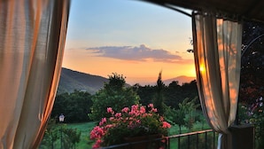 A sunset view from a balcony with curtains, a garden, and a distant mountain range.