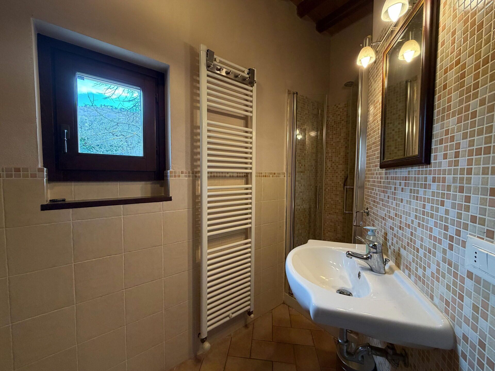 Ein Badezimmer mit Fliesenboden, einem weißen Waschbecken, einer Duschkabine aus Glas, einem Handtuchhalter und einem Fenster mit Blick auf die Landschaft.