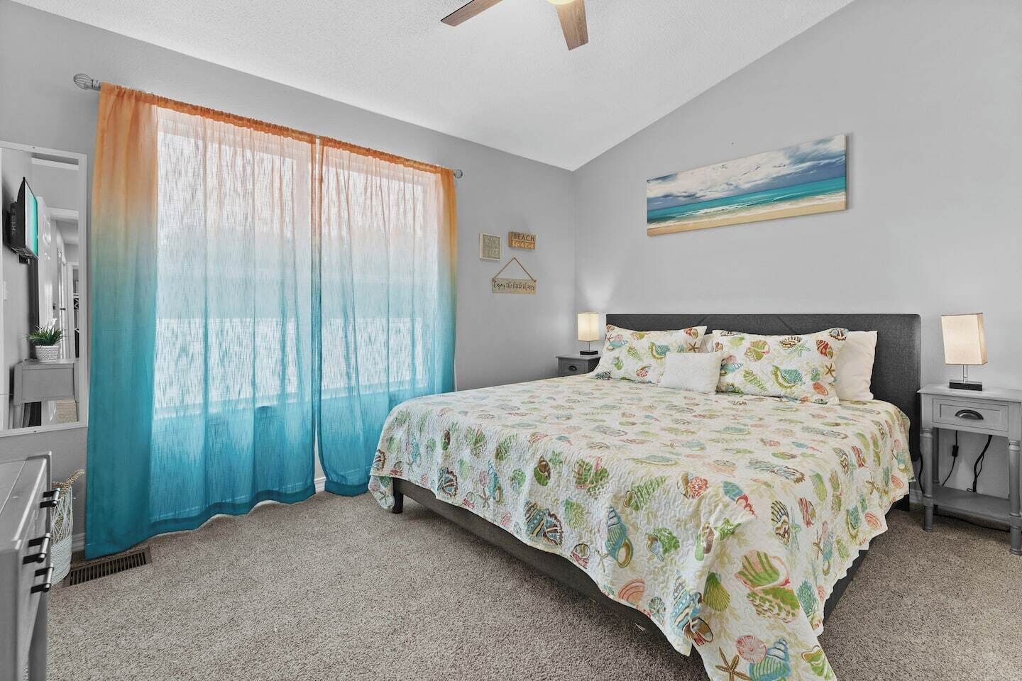 A bedroom with a bed, two bedside tables, a ceiling fan, a window with sheer curtains, and a beach-themed painting on the wall.