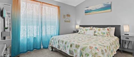 A bedroom with a bed, two bedside tables, a ceiling fan, a window with sheer curtains, and a beach-themed painting on the wall.