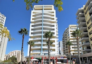 A modern multi-story hotel with balconies, palm trees, and a clear blue sky.