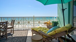 Ein Balkon mit Blick auf den Strand, ein Holzstuhl, eine gepolsterte Liege und ein kleiner runder Tisch.