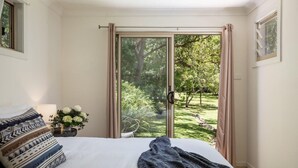 Un dormitorio con cama, ventana con cortinas, vista a un jardín y una maceta con flores.