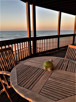 Une terrasse en bois avec une table ronde et des chaises, donnant sur une plage et l’océan.