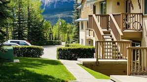 A two-story wooden building with a balcony, a driveway, and a mountain backdrop. - PRIVATE Entrance, Mountain View ,2 BR/1.5 BT  (Harvie Heights)
