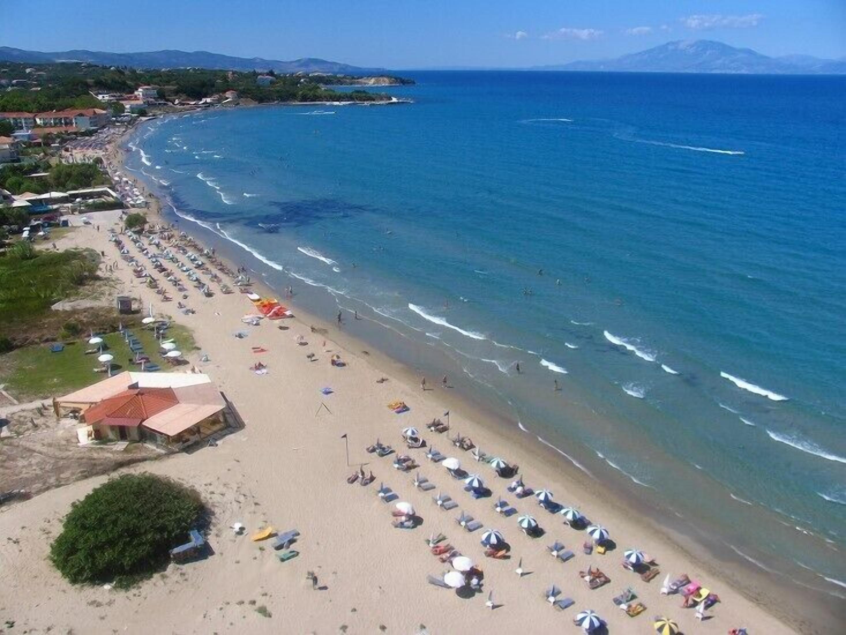 Una playa con filas de tumbonas y sombrillas, un pequeño edificio y una vista despejada del mar y montañas distantes.