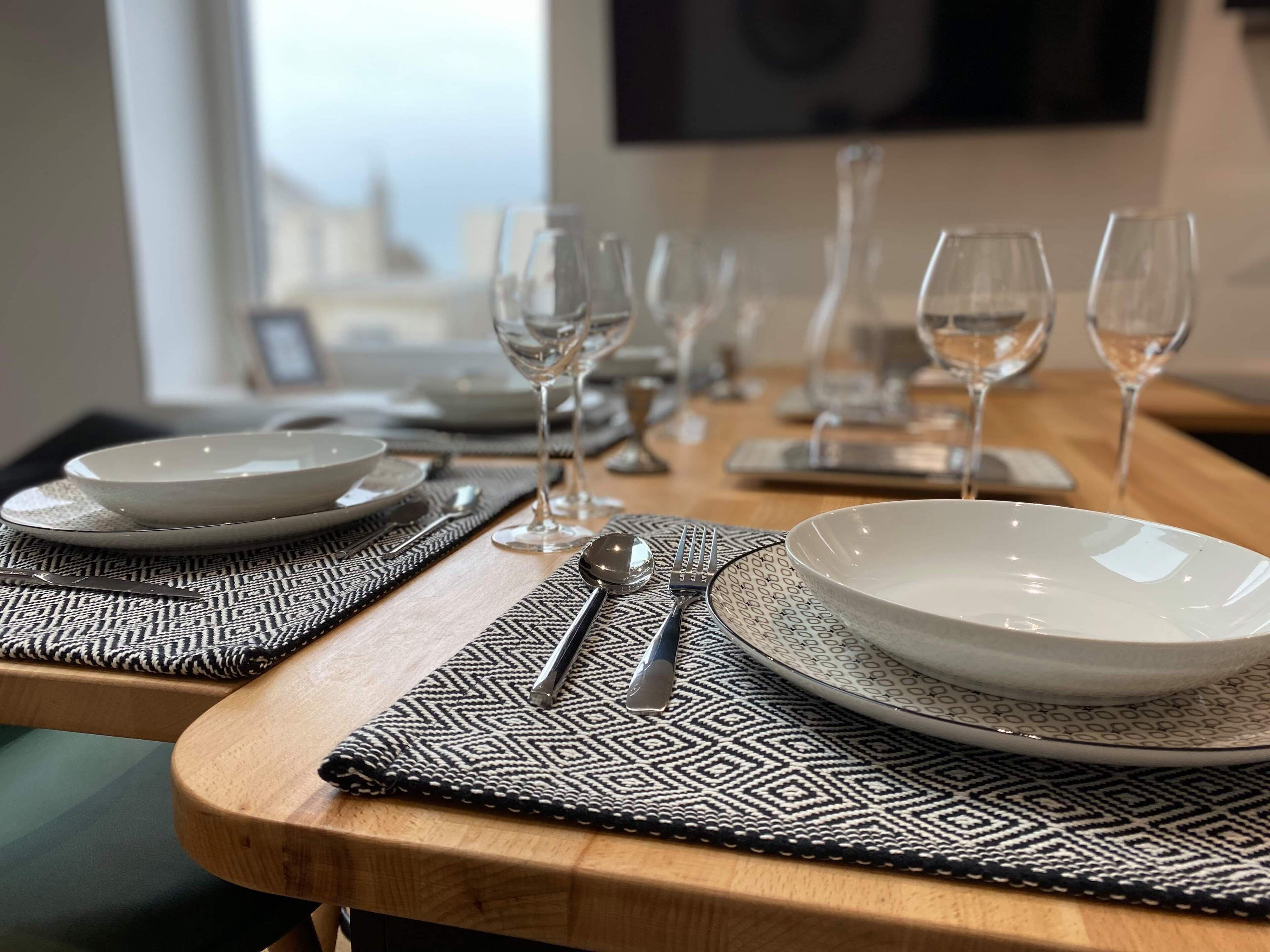 A dining table set with plates, glasses, and cutlery, with a patterned placemat and a view of the cityscape through the window.