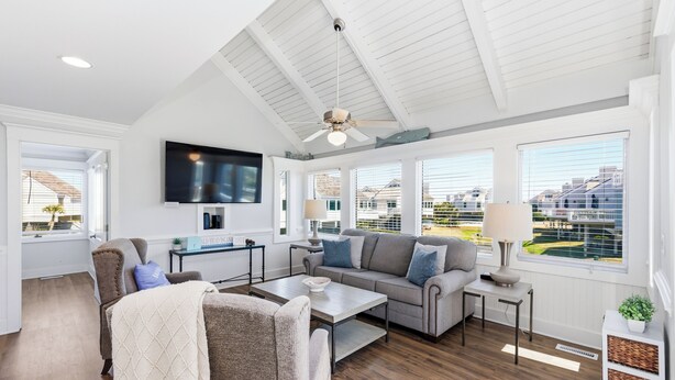 Un vaste salon doté d’un ventilateur de plafond, d’un téléviseur à écran plat, d’un canapé gris, d’une table basse et de grandes fenêtres offrant une vue sur le quartier.