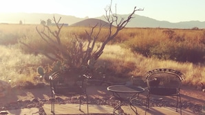 Deux chaises en métal et une table sont disposées sur une terrasse en pierre, offrant une vue sur un champ aride et herbeux, avec des montagnes en arrière-plan.