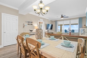 A dining area with a wooden table, chairs, a chandelier, a television, and a view of the ocean through large windows.