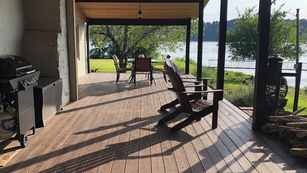 Een overdekt terras met een barbecue, stoelen en uitzicht op een meer.