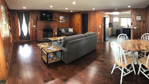 Een houten interieur met een bank, een eettafel, stoelen, een open haard en een keukenhoek.