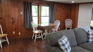 Een kamer met houten wanden, een grijze bank met geruite kussens, een houten eettafel met stoelen en een raam met gordijnen.