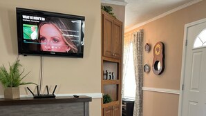 A living room with a flat-screen TV mounted on the wall, a fireplace, a wooden cabinet, and a door leading to another room. - Great property for contract healthcare workers! (Hamlet)