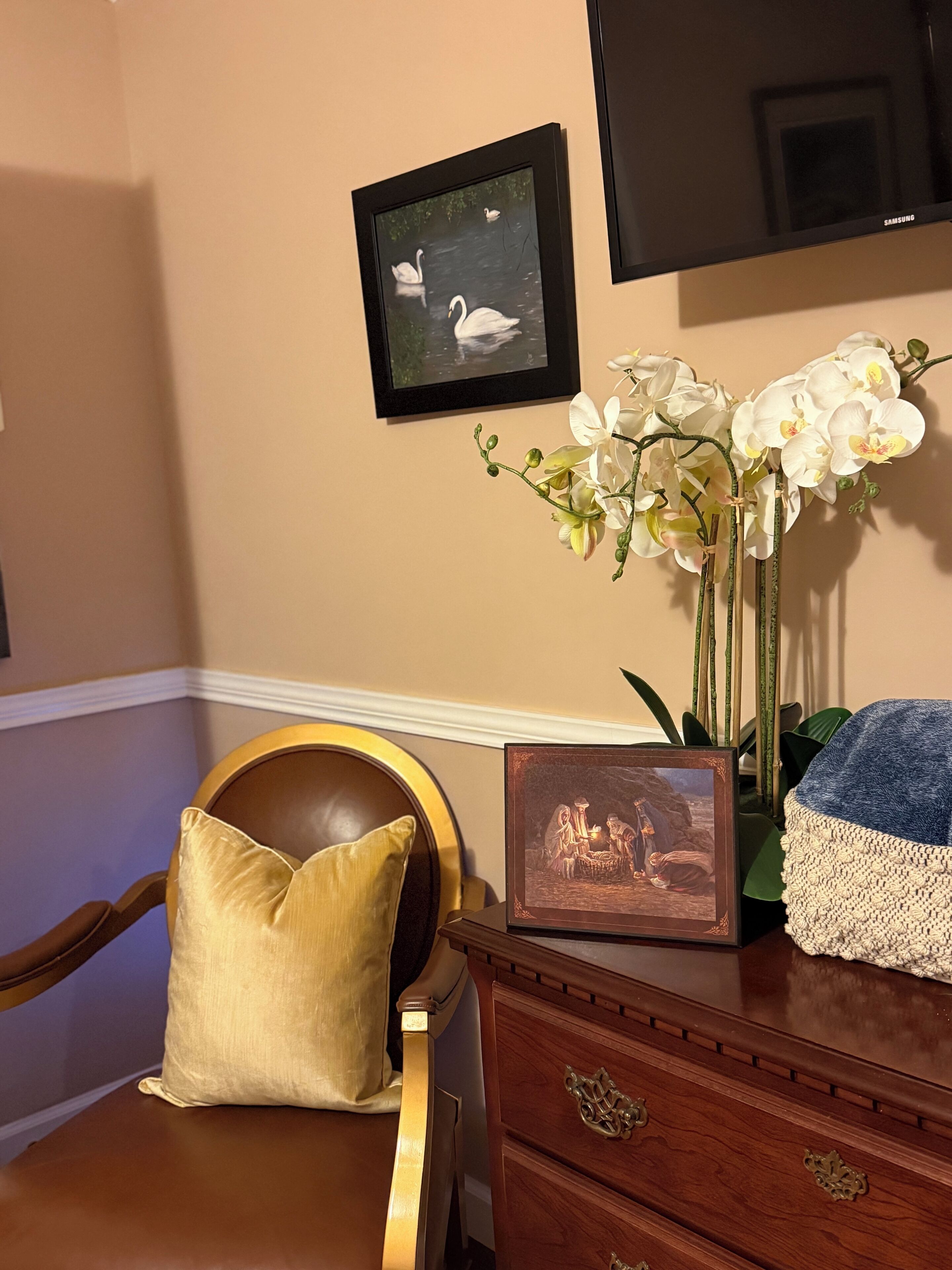 A room with a wooden chair, a cushion, a framed picture, a vase of orchids, and a television on the wall.