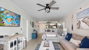 A living room with a television, a ceiling fan, a sofa, a coffee table, and a kitchen in the background. - Ground floor easy access! A127 (Fort Walton Beach)