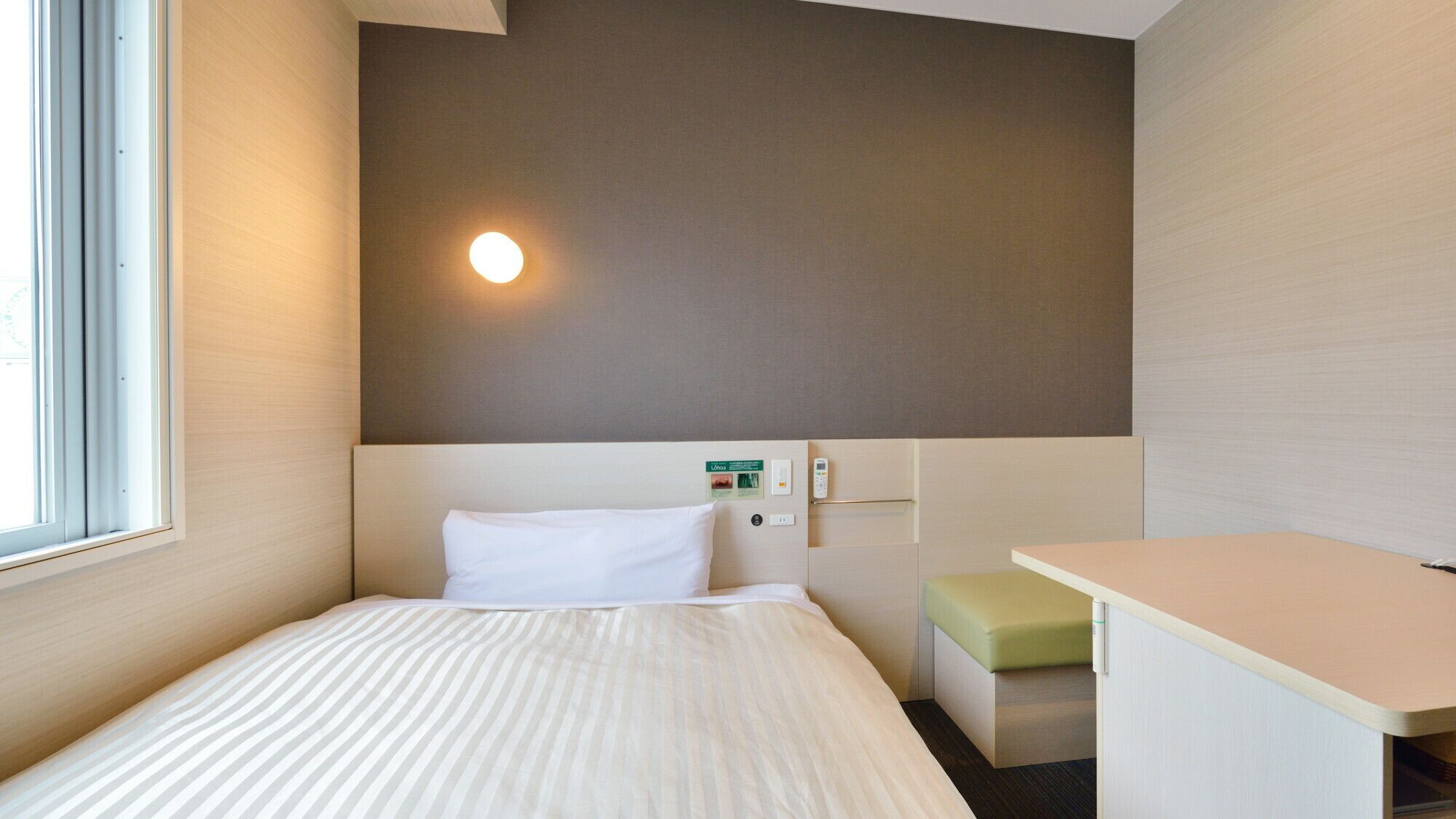 A hotel room with a bed, a desk, a small bench, and a wall-mounted control panel.