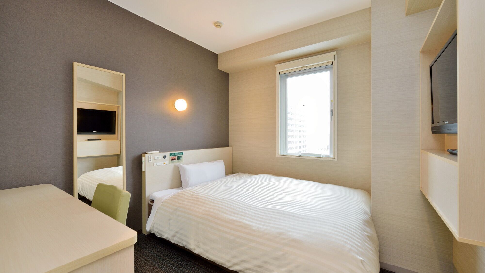 A hotel room with a bed, a desk, a TV mounted on the wall, and a window.