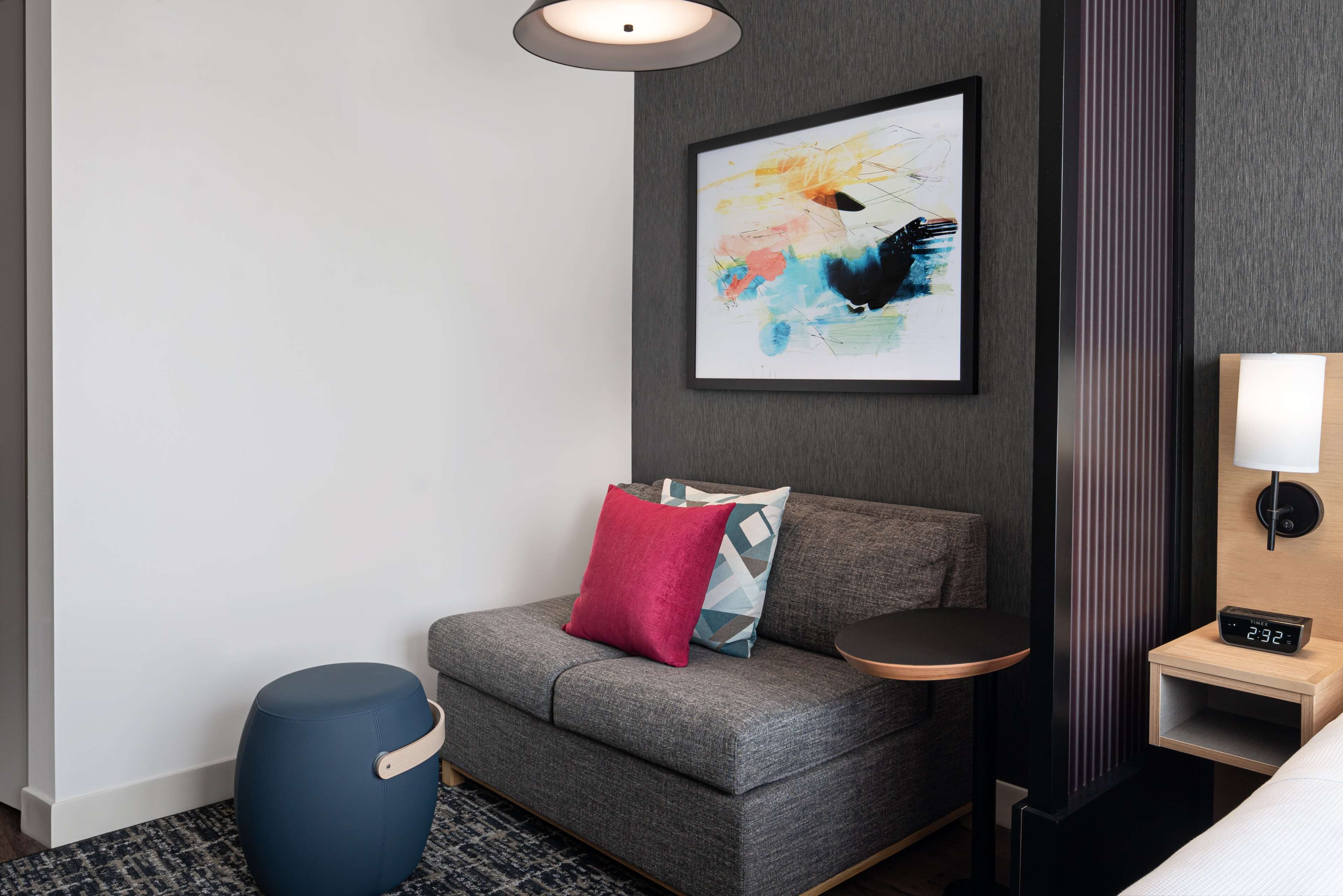 A modern hotel room with a grey sofa, a blue ottoman, a small round table, a wall-mounted lamp, and a framed abstract painting.