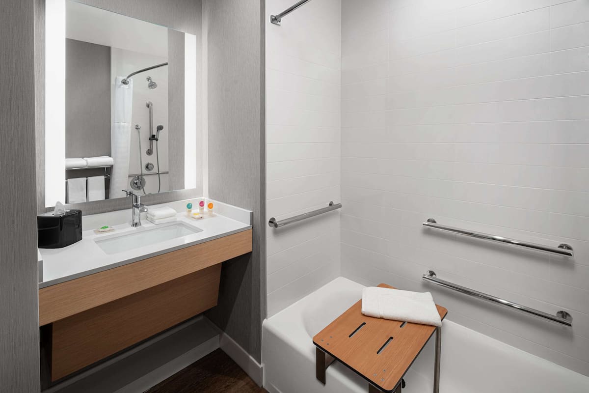 A modern bathroom with a white sink, mirror, and shower. It includes a wooden vanity, a small wooden stool, and grab bars on the walls.