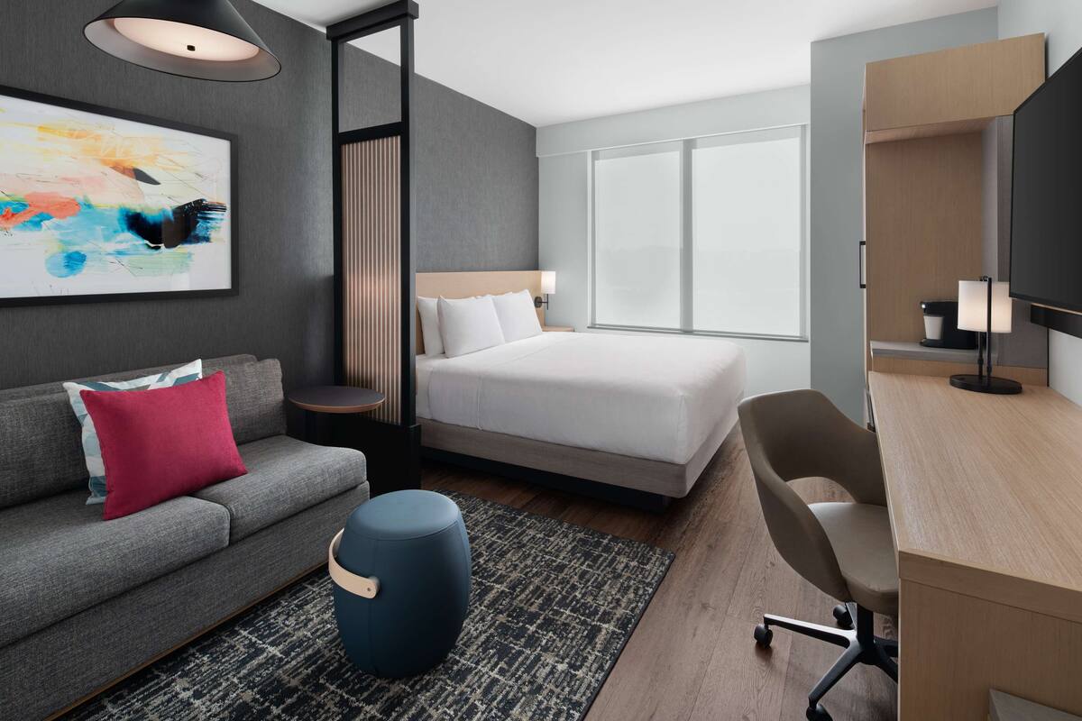 A modern hotel room with a bed, a sofa, a desk, and a large window.