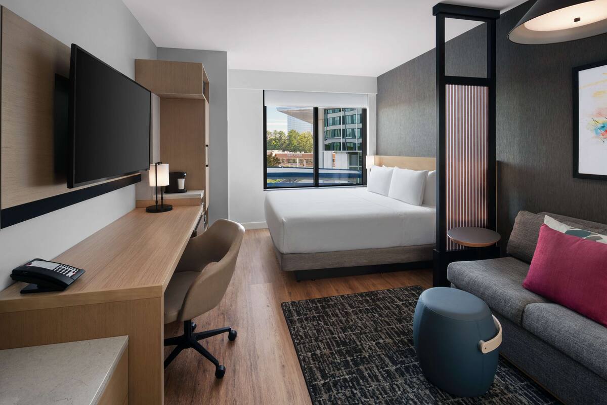 A hotel room with a bed, a desk with a computer, a TV, a sofa, and a view of a building and trees.
