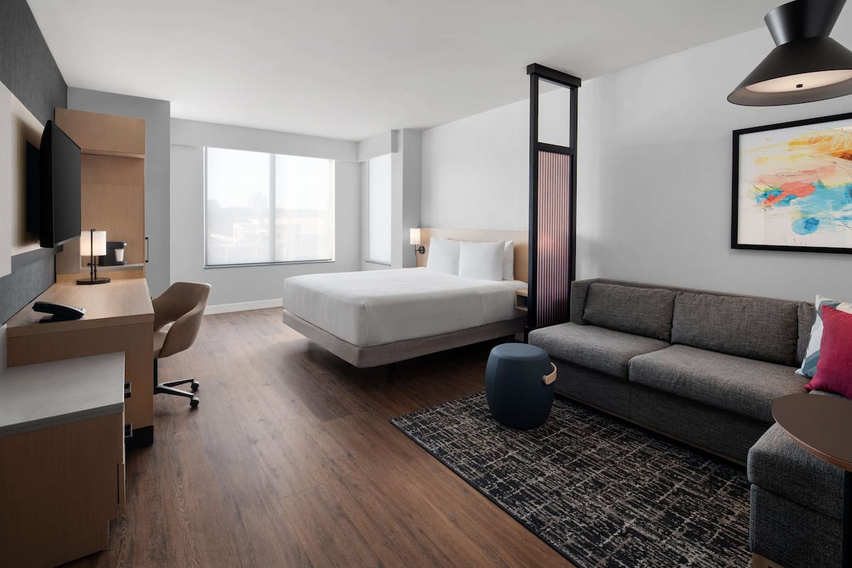 A modern hotel room with a bed, a sofa, a desk with a chair, a TV, and a large window.