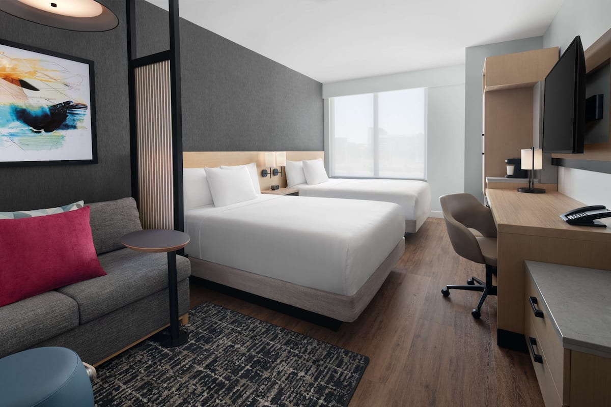 A modern hotel room with two beds, a desk, a chair, a TV, and a large window.