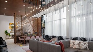 A modern living area with a sectional sofa, dining table, and chairs. There are hanging lights and large windows with sheer curtains. - Hyatt Place Atlanta / Perimeter Center (Atlanta)