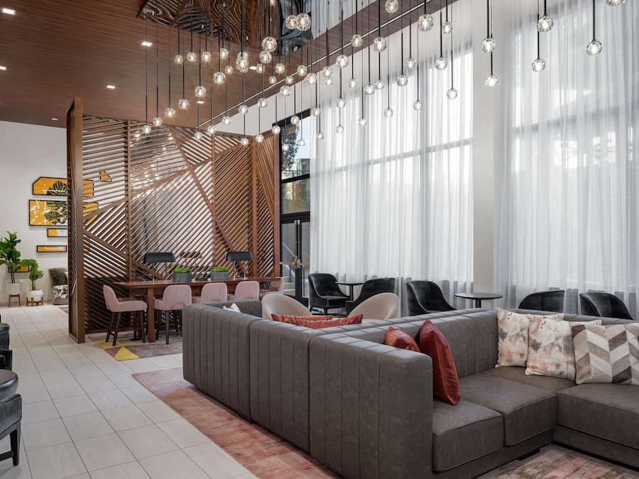 A modern living area with a sectional sofa, dining table, and chairs. There are hanging lights and large windows with sheer curtains.