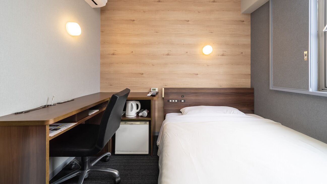 A hotel room with a wooden headboard, a desk with a chair, a bed, and a small kitchenette area.