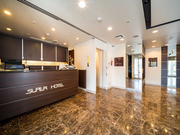 Zona de recepción de un hotel con piso de mármol, mostrador de recepción identificado con el rótulo "Super Hotel" y una vista despejada del vestíbulo.