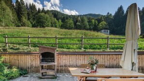 An outdoor patio with a stone barbecue grill, wooden table, and chairs, set against a backdrop of a wooden fence and a forested hillside.