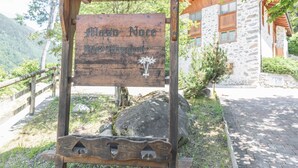 A wooden sign with 'Maso Noce' and 'Haus Maggimitt' written on it, mounted on a wooden structure with a stone building in the background.