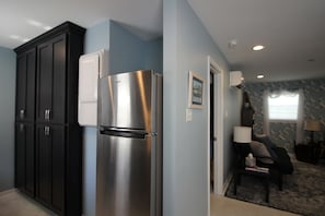 Enjoy cooking in this well-equipped kitchen with a refrigerator and dark cabinets, adjacent to a cozy living area with a rug, lamp, and picture.