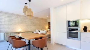 A modern kitchen with a built-in oven and a dining area with a wooden table and brown chairs. - Tropical View Cascais - CheckinHome (Guincho)