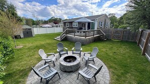 A backyard with a fire pit, outdoor seating, a wooden deck, and a house with a covered porch. - Clean and bright beach cottage, walking distance to all. (Yarmouth)